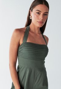 Groene halterjurk met een geplooid borststukontwerp, gestructureerde stof en een aansluitende taille die uitloopt in een uitlopende rok.