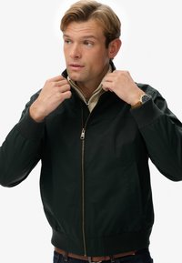 Chaqueta bomber negra hecha de tejido sintético suave, con cierre de cremallera, puños acanalados y bolsillos laterales, llevada sobre una camisa beige.