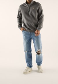 Sudadera gris con cremallera y un pequeño logo dorado, combinada con jeans desgastados en azul claro. Calzado: zapatillas beige.