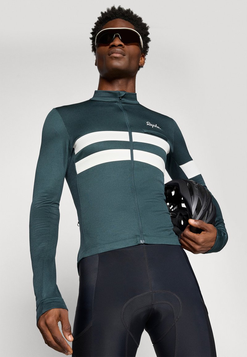Dark green cycling jersey with white stripes, long sleeves, and a zipper. Black cycling shorts, helmet held in hand, minimalist design.