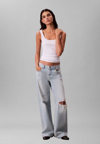 Débardeur blanc cintré avec un col en U, associé à un jean large bleu clair avec des détails usés. Des chaussures foncées complètent le look.