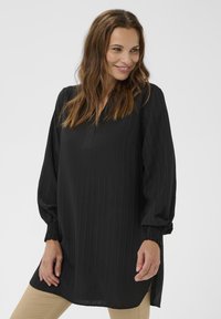 Žena s dlhými hnedými vlasmi, oblečená v voľnej čiernej tunike s dlhým rukávom, s textúrovanými vertikálnymi pruhmi a béžovými nohavicami, usmievajúca sa a pozerajúca sa nabok.