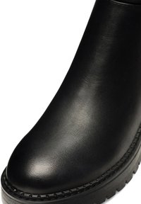 Bota de cuero negro con punta redondeada y suela gruesa con dibujo, que presenta costuras visibles a lo largo del borde inferior.