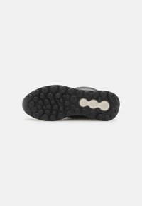 Suela de zapatilla negra con un agarre patternado, que presenta texturas circulares y hexagonales. Incluye un elemento de amortiguación blanco en el centro.