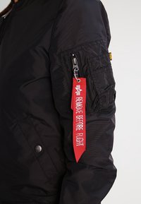 Zwarte bomberjack met een gestructureerde afwerking, voorzien van een zak op de mouw met rits en een rood stoffen label met de tekst "REMOVE BEFORE FLIGHT".