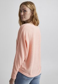 Textuurtrui in roze met een brede halslijn, afgeknipte schouders en lange mouwen, gecombineerd met een blauwe spijkerbroek tegen een neutrale achtergrond.