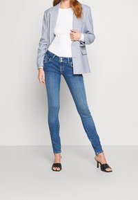 Modré skinny džíny s designem pěti kapes, spárované s bílým žebrovaným topem a světlým kostkovaným blejzrem.  Černé sandály na podpatku dokončují vzhled.