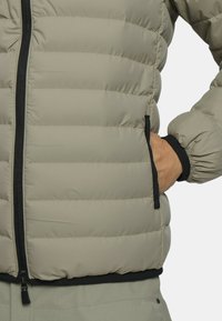 Beige Pufferjacke mit Reißverschluss auf der Vorderseite. Horizontal gestepptes Design, elastische Bündchen und Seitentaschen. Dezenter Glanz auf der Stoffoberfläche.