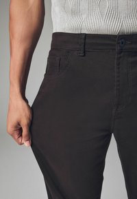Dark brown pants with a fitted design, made of soft fabric. Features a front pocket and a visible button closure at the waistband.