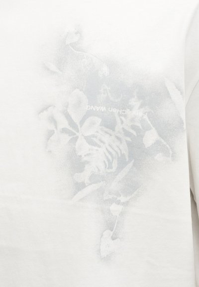 Λευκό ύφασμα με αχνό, λουλουδάτο και τίγρε μοτίβο σε ανοιχτό γκρι, συνοδευόμενο από μερικώς ορατό ανάποδο κείμενο.