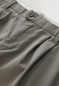 Gros plan sur la ceinture d’un pantalon gris avec fermeture par bouton, passants de ceinture et détail plissé à l’avant.