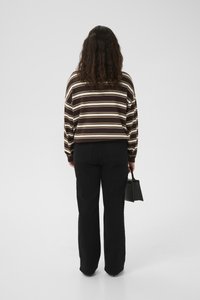 Woman with long curly hair wearing a brown striped sweater and black pants, holding a small black handbag, standing against white background.