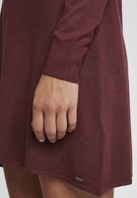 Robe tricotée bordeaux à manches longues, avec des poignets côtelés et un ourlet légèrement évasé. Petite étiquette en métal sur le côté inférieur. Texture douce.