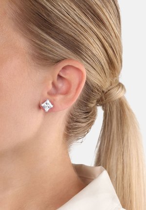 Capelli biondi raccolti, indossa un orecchino a cerchio quadrato in argento all'orecchio sinistro, su uno sfondo chiaro e semplice.