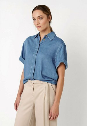 Woman with brown hair tied back, wearing a blue short-sleeve button-up shirt and high-waisted beige pleated trousers.