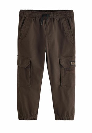 Dreng iført en lys grå oversized t-shirt, mørke cargobukser med elastiske manchetter og hvide sneakers, stående mod en ensfarvet beige baggrund.