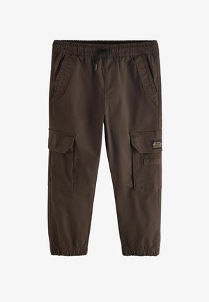 Dreng iført en lys grå oversized t-shirt, mørke cargobukser med elastiske manchetter og hvide sneakers, stående mod en ensfarvet beige baggrund.
