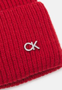 Piros kötött sapka bordázott textúrával, ezüst "CK" logó emblémával az elején, kiemelve a dizájn és anyag részleteit.