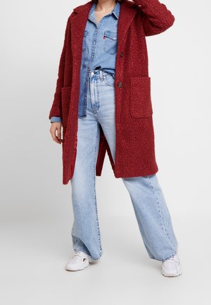 Femme portant un long manteau rouge texturé sur une chemise en jean et un jean bleu clair, posant avec une main touchant la tête.