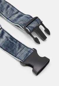 Sangle en tissu motif jean avec boucle en plastique noir à déclenchement latéral partiellement déconnectée sur fond blanc.
