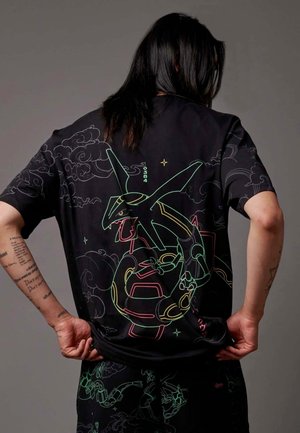 Persona con capelli lunghi e scuri che indossa una maglietta nera con un design di drago robotico in stile neon e pantaloni neri coordinati con motivi simili.