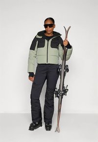 Šviesiai žalia ir juoda paminkštinta striukė su užtrauktuku; derinama su juodomis slidinėjimo kelnėmis. Modelis laiko slides, nešioja akinius nuo saulės ir juodus batus.