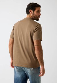 Brązowy t-shirt z okrągłym dekoltem wykonany z miękkiego materiału, z krótkimi rękawami i luźnym krojem, zestawiony z jasnoniebieskimi dżinsami i widocznym logo na pasku.