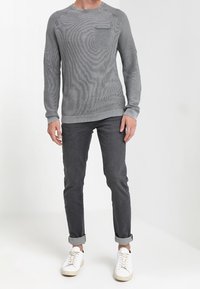 Grauer gestrickter Pullover mit langen Ärmeln, Rundhalsausschnitt und einer Brusttasche, kombiniert mit dunklen Jeans und weißen Sneakers. Glatte Textur.