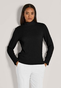 Frau mit langen dunklen Haaren trägt einen schwarzen strukturierten Rollkragenpullover und weiße Hose, Hände in den Taschen, neutraler Hintergrund.