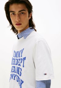 Hvid bomulds T-shirt med blå grafisk tekst; lagd oven på en blå og hvid ternet skjorte med krave. Har brandlogo på ærmet.