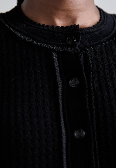 Lähikuva henkilöstä, joka pukeutuu mustaan teksturoituun neulecardiganiin, jossa on mustat napit ja koristeellinen viimeistely kauluksessa ja napituslinjalla.