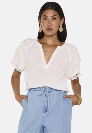 Donna con una camicetta bianca con maniche di pizzo, pantaloni a vita alta azzurri chiaro, orecchini ovali e braccialetti, con le mani in tasca.