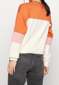 Žena oděná v barevném svetru s bloky oranžové, bílé a růžové, doplněná o vysokým pasem tmavě šedé džíny, zobrazena zezadu.