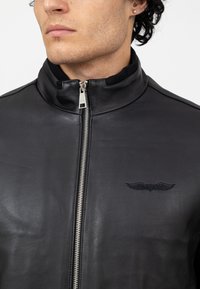 Giacca di pelle nera con colletto alto, zip argentata e logo ricamato a forma di ala sul lato sinistro. Tessuto liscio e vestibilità su misura.