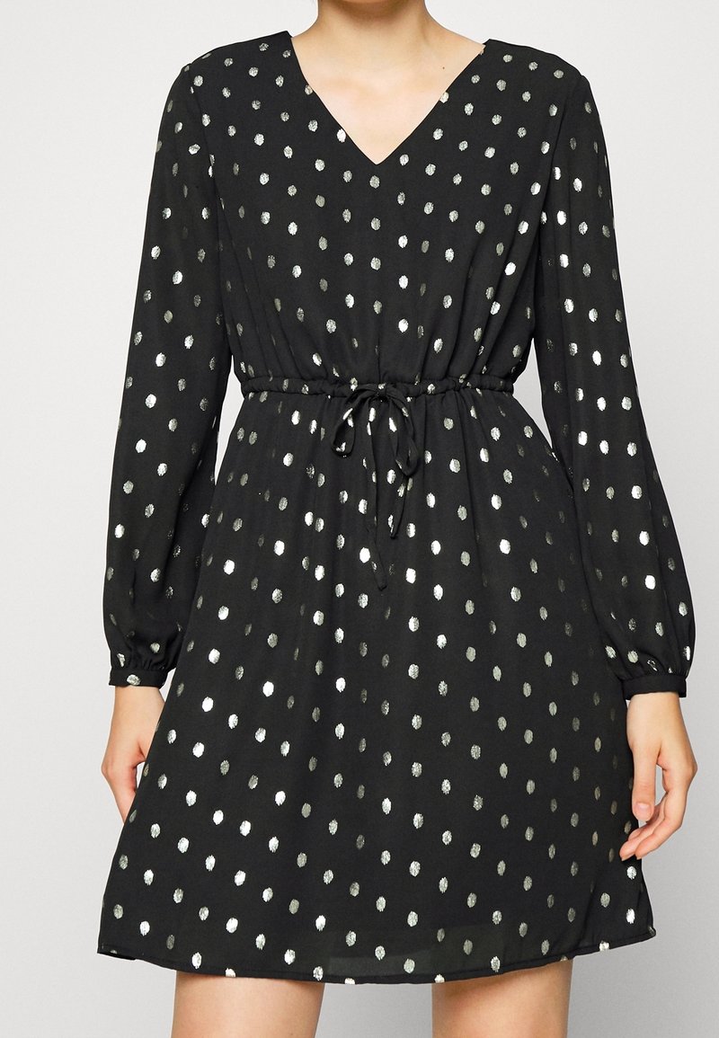 Robe noire à manches longues avec des pois argentés, col en V et lien ajustable à la taille portée par une personne du cou jusqu'à mi-cuisse.