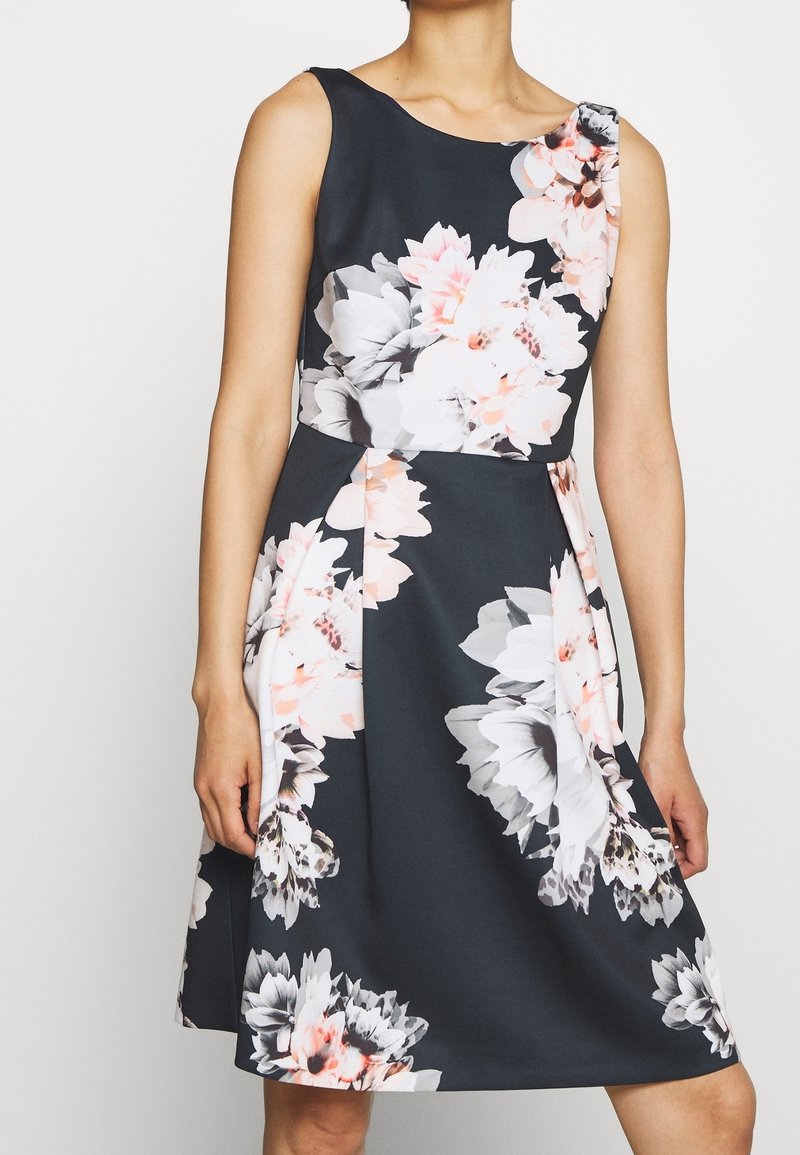 Robe sombre sans manches avec de grands imprimés floraux blancs et roses, taille cintrée et jupe évasée portée par une personne aux bras détendus.