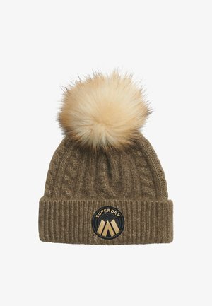 Bruine cable-knit muts met namaakbont pompon bovenop, voorzien van een ronde zwart-gouden "SUPERDRY" logo patch op de omgeslagen band.
