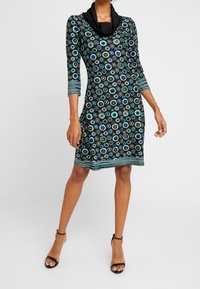 Czarna sukienka z dekoltem w kształcie łezki, rękawami o długości 3/4 i wzorem kolorowych koncentrycznych kół. Paski na dolnym brzegu i dopasowany krój.