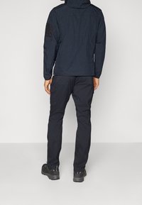 Man som bär en svart huvtröja och svarta cargo-byxor, står vänd mot en vit vägg med händerna avslappnat vid sidorna.