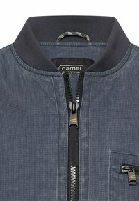 Nahaufnahme des Kragens einer blau-grauen Reißverschlussjacke mit schwarzem, geripptem Besatz und einer kleinen Reißverschlusstasche auf der Brust.