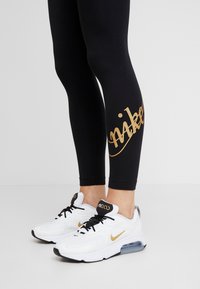 Svarta leggings med en guldig "Nike"-logotyp på vaden, kombinerat med vita sneakers med svarta detaljer och en synlig Air-enhet.