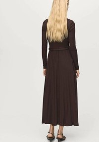 Donna con lunghi capelli biondi che indossa un vestito attillato di colore marrone scuro a maniche lunghe con gonna pieghettata e tacchi neri con cinturini, rivolta di spalle.