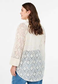 Bluzka koronkowa w jasnym kremie o półprzejrzystej strukturze, z rękawami o długości 3/4 i zaokrąglonym dołem, ozdobiona kwiatowym wzorem koronkowym.