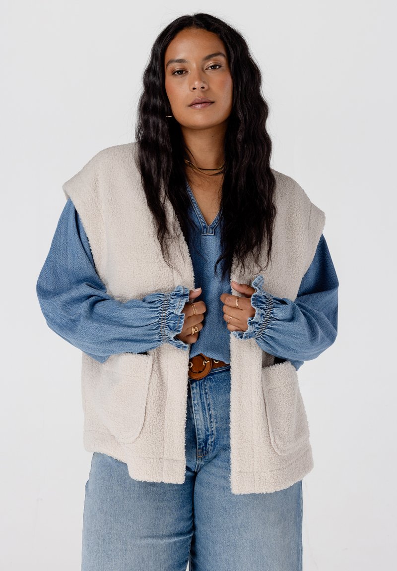 Beige fleece vest met grote zakken, gelaagd over een blauwe spijkerblouse. Zachte textuur, open voorkant en een licht oversized ontwerp.