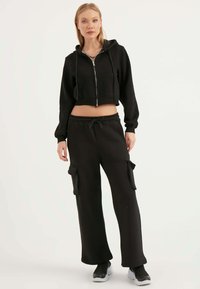Hoodie cropped nero con zip frontale e coulisse regolabile, abbinato a pantaloni cargo neri a vestibilità ampia con tasche laterali.