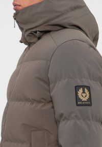 Brun pufferjakke med høj krave og lynlås. Har et Belstaff-logo-patch på venstre ærme og en quiltet tekstur.