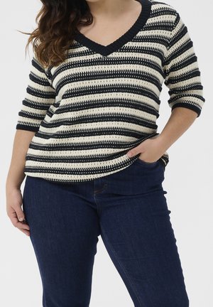 Personne portant un pull à rayures noires et blanches avec un col en V et des manches mi-longues, ainsi qu'un jean en denim bleu foncé, la main dans la poche.