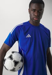 Blå idrettsdrakt med hvite detaljer og tre-stripe merkevare, holdt sammen med en svart-hvite mønstrete fotball mot en nøytral bakgrunn.