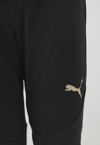Svarta sportshorts i slät tyg med en guld Puma-logga. Designen inkluderar sidofickor och en strukturerad panel på benen.