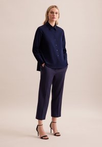 Granatowa koszula zapinana na guziki z długim rękawem, gładki materiał; w połączeniu z granatowymi spodniami 3/4 i czarnymi sandałkami na obcasie, minimalistyczny design.